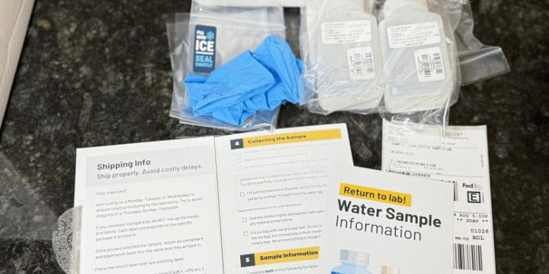 PFAS water testing kit with sampling bottles, protective gloves, instructions, and return-to-lab shipping label on a kitchen counter.